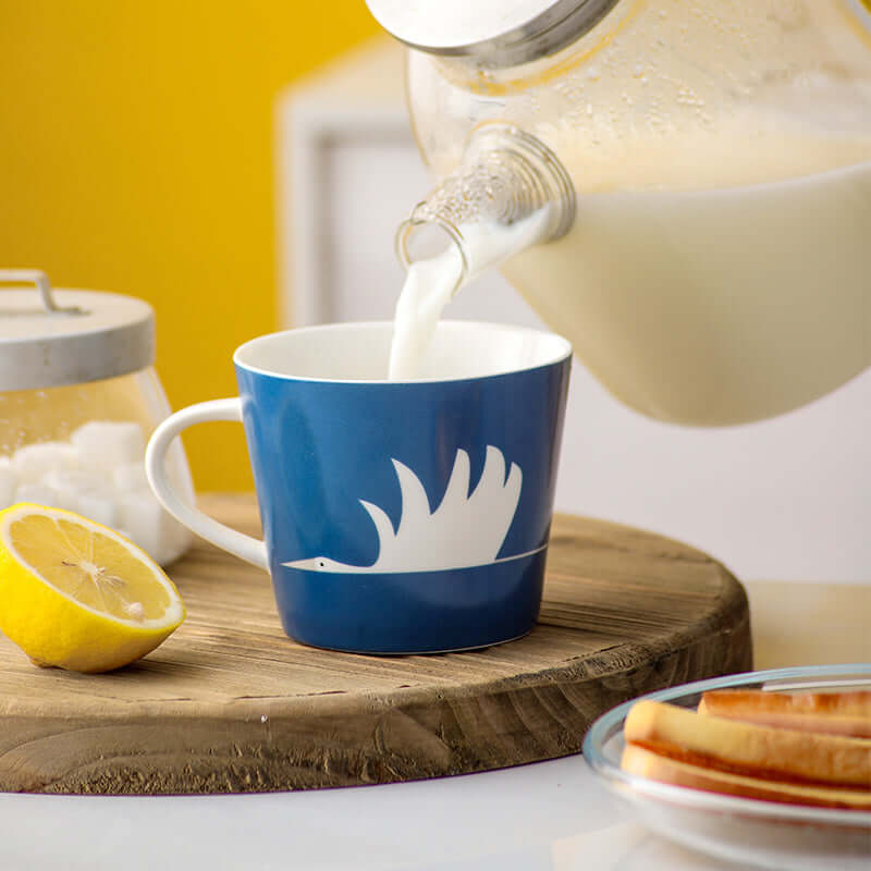 Blue ceramic mug with swan design being filled with milk, placed on a wooden board with lemon and sugar jar in a cozy setting.