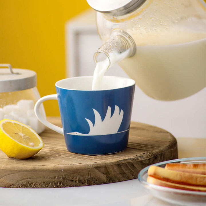 Blue ceramic mug with swan design being filled with milk, placed on a wooden board with lemon and sugar jar in a cozy setting.