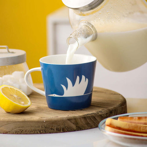 Blue ceramic mug with swan design being filled with milk, placed on a wooden board with lemon and sugar jar in a cozy setting.